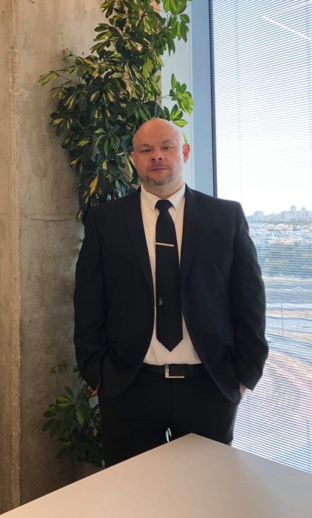 תמונתו של עורך דין יגאל וסיליאב, לבוש בחליפה שחורה ועניבה - מדי משפט, העומד לצד חלון וליד שולחן ועציץ
